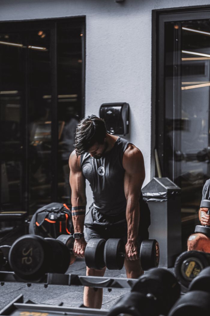 Elementor #1456 A muscular man in a tank top lifts heavy dumbbells in a gym setting.
